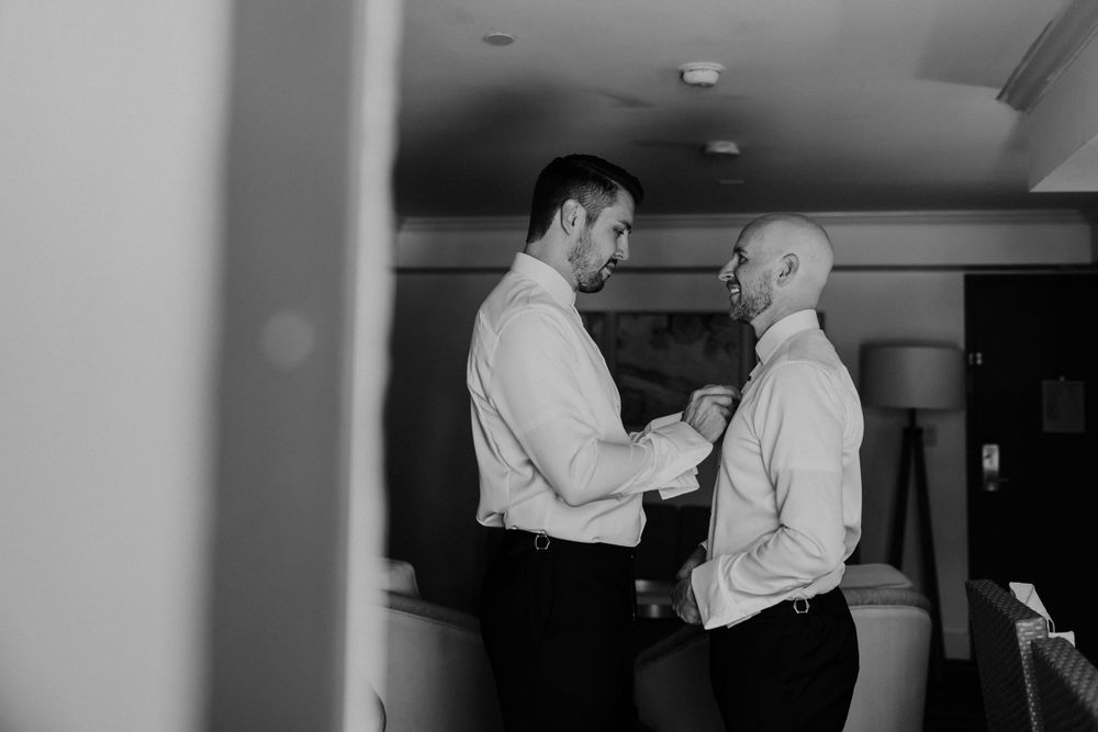Two people in white dress shirts share an intimate moment while adjusting clothing before a formal event.