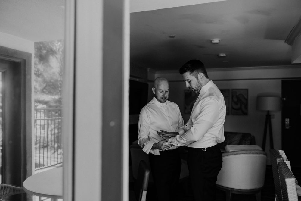 Two people in white dress shirts helping each other get ready in a room with natural lighting.