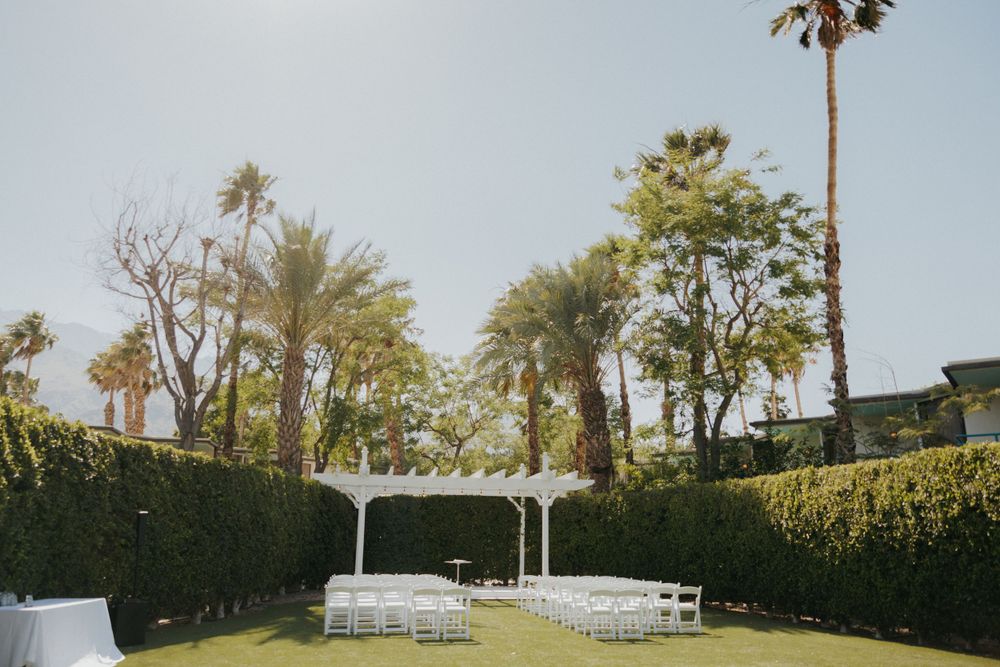 Outdoor wedding venue with white pergola and chairs surrounded by manicured hedges and palm trees.