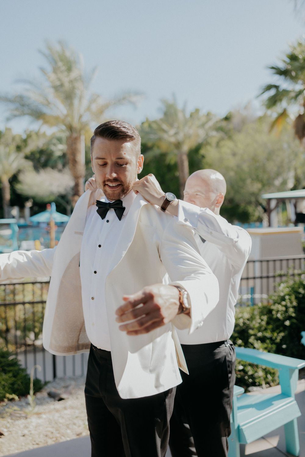 Two people in formal white tuxedos getting ready outdoors by a pool with palm trees.