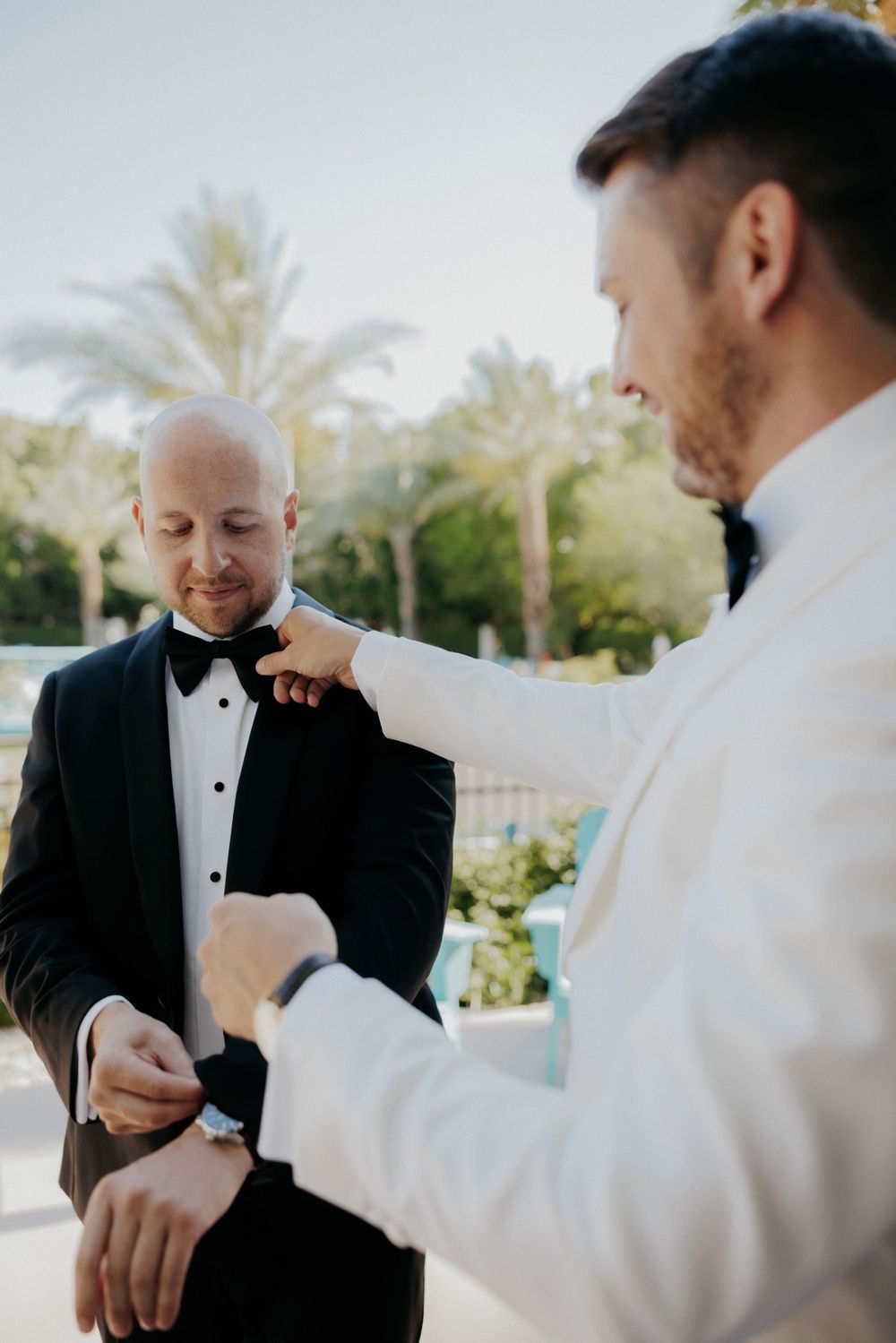 Two people in tuxedos help each other with final adjustments to their formal attire.