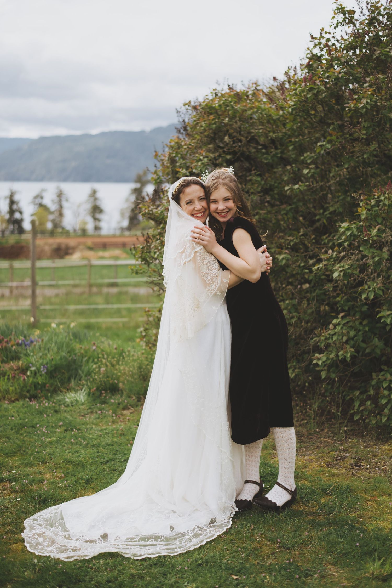 Two people embrace joyfully outdoors, one in a white wedding dress and one in a black dress.