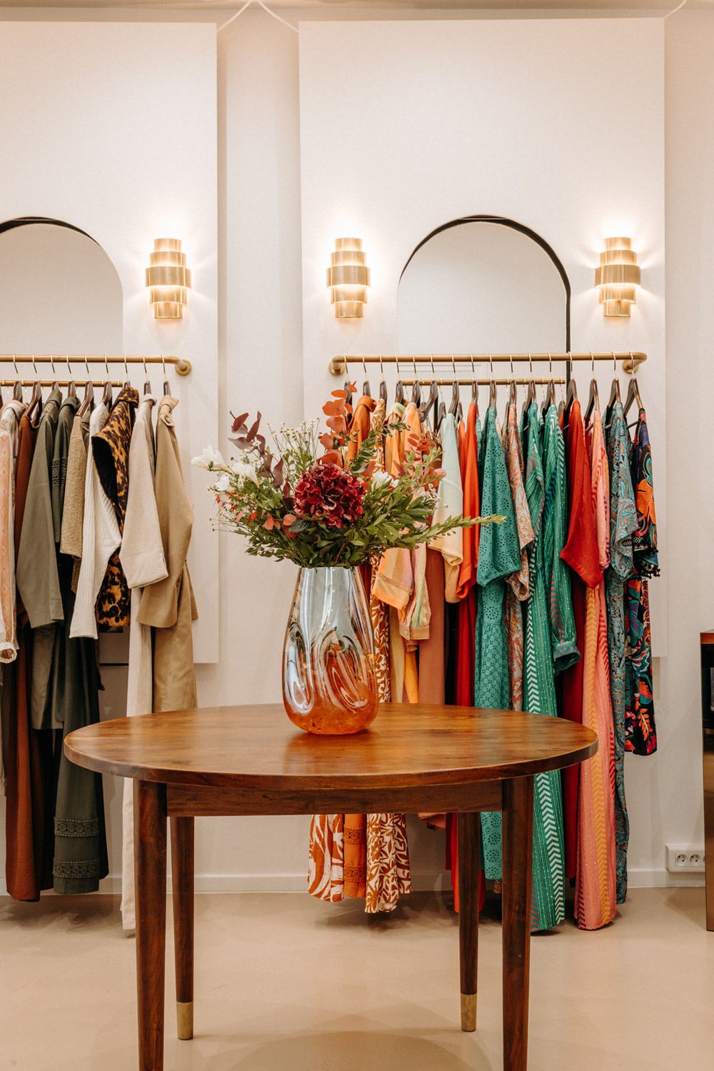 Elegant boutique clothing store with colorful garments on gold racks, arched mirrors, wall sconces, and a wooden table with floral vase.