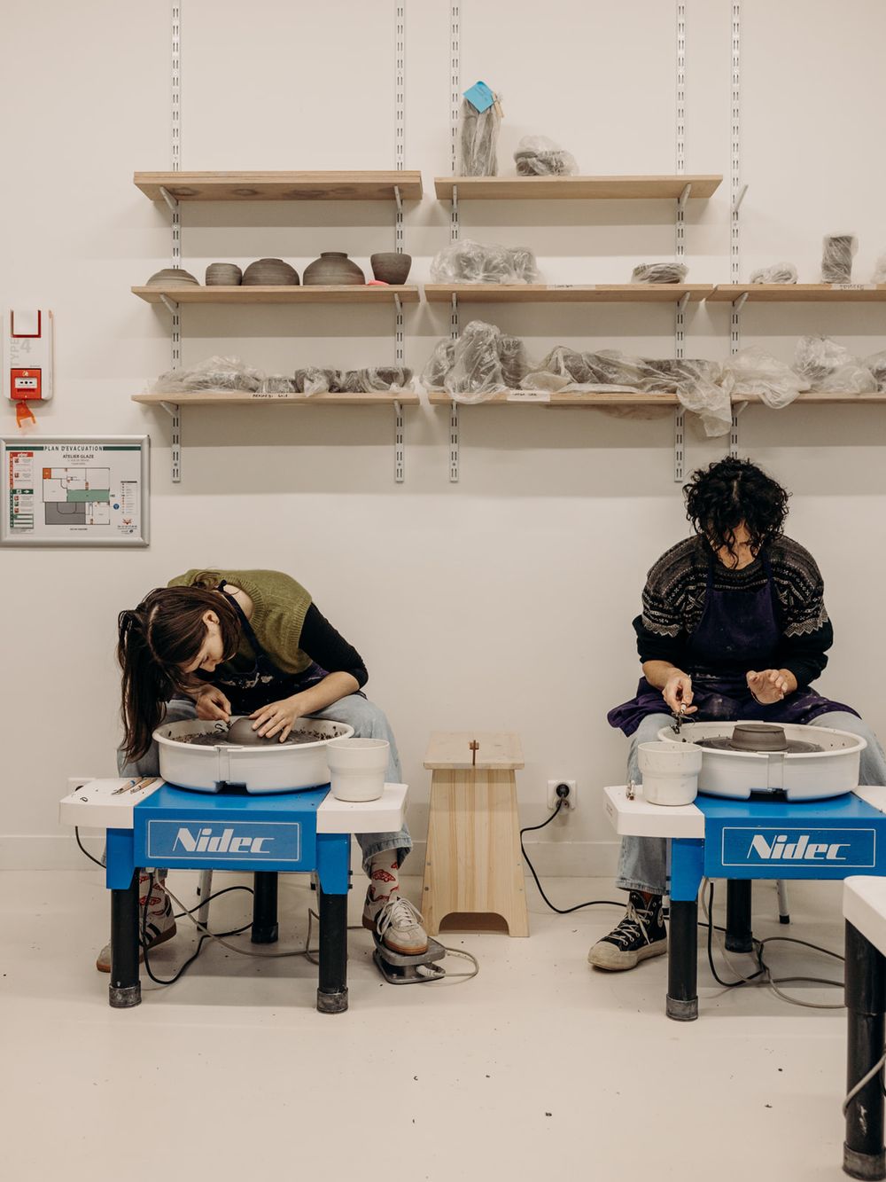 Two people using Nidec pottery wheels in a bright ceramic studio with clay works displayed on shelves above.
