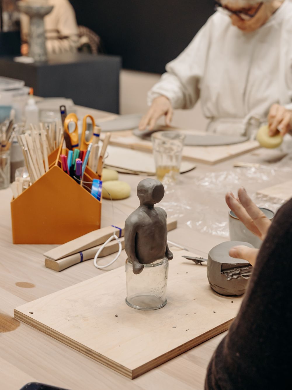 Clay sculpture of a small figure sits on a glass jar at an art workshop table with craft supplies and students working.