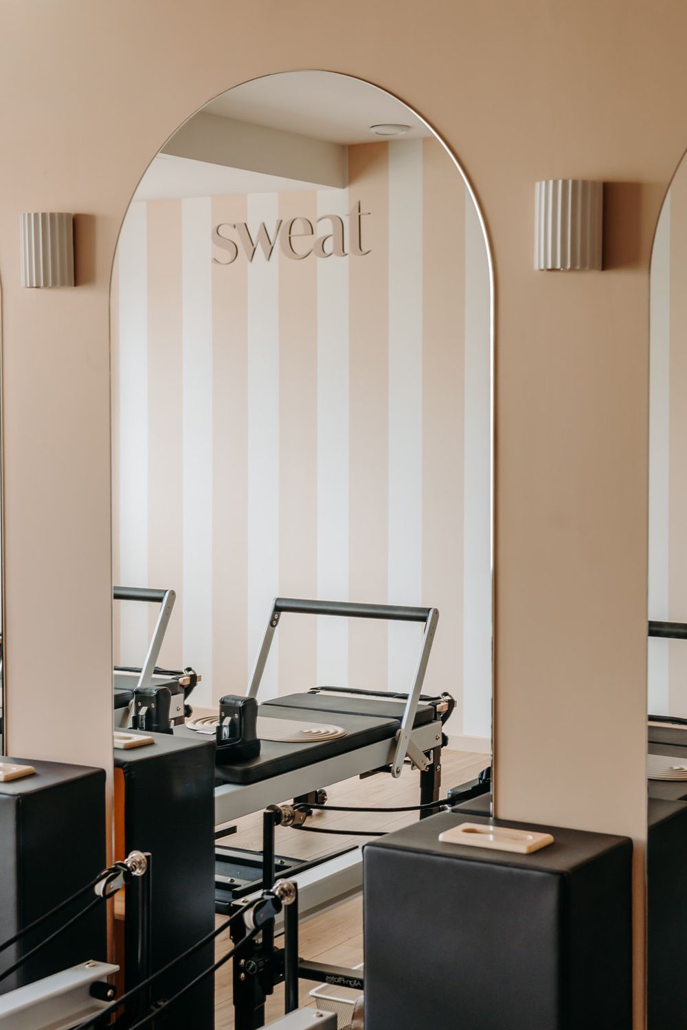 Elegant Pilates reformer studio with arch mirror, 'sweat' signage, striped walls, and modern black equipment.