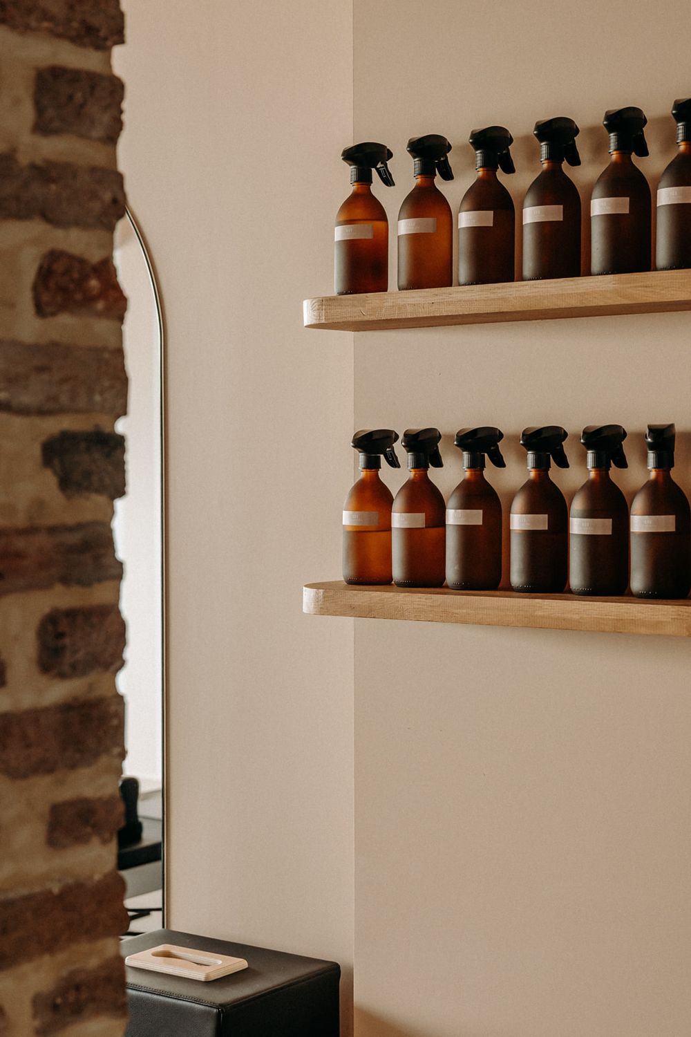 Amber spray bottles with white labels displayed on wooden shelves against a cream wall near a brick archway.