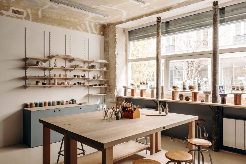 Bright pottery studio with wooden worktable, wall-mounted shelves of ceramics, and large windows with potted plants.