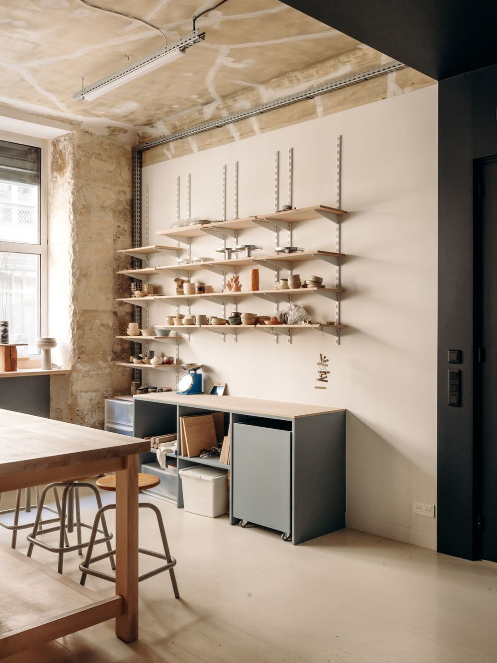 Industrial workspace with wall-mounted shelves displaying ceramics, a wooden worktable, metal stools, and modular storage units.