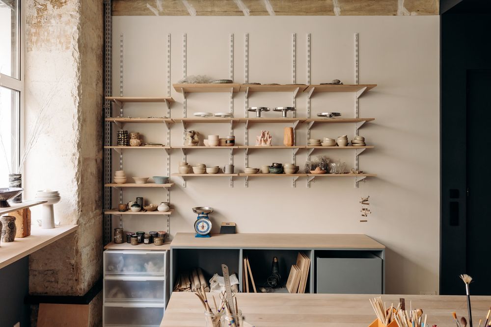 Rustic pottery studio with wooden shelves displaying ceramic pieces above storage cabinets and a workbench.