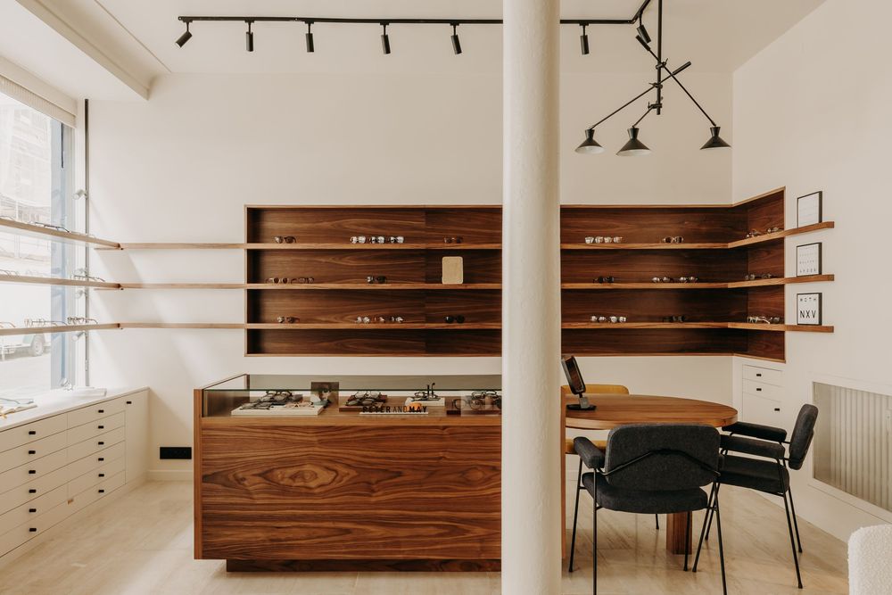 Modern optical shop interior with walnut wood display cases, shelving with eyewear, and minimalist Scandinavian design.