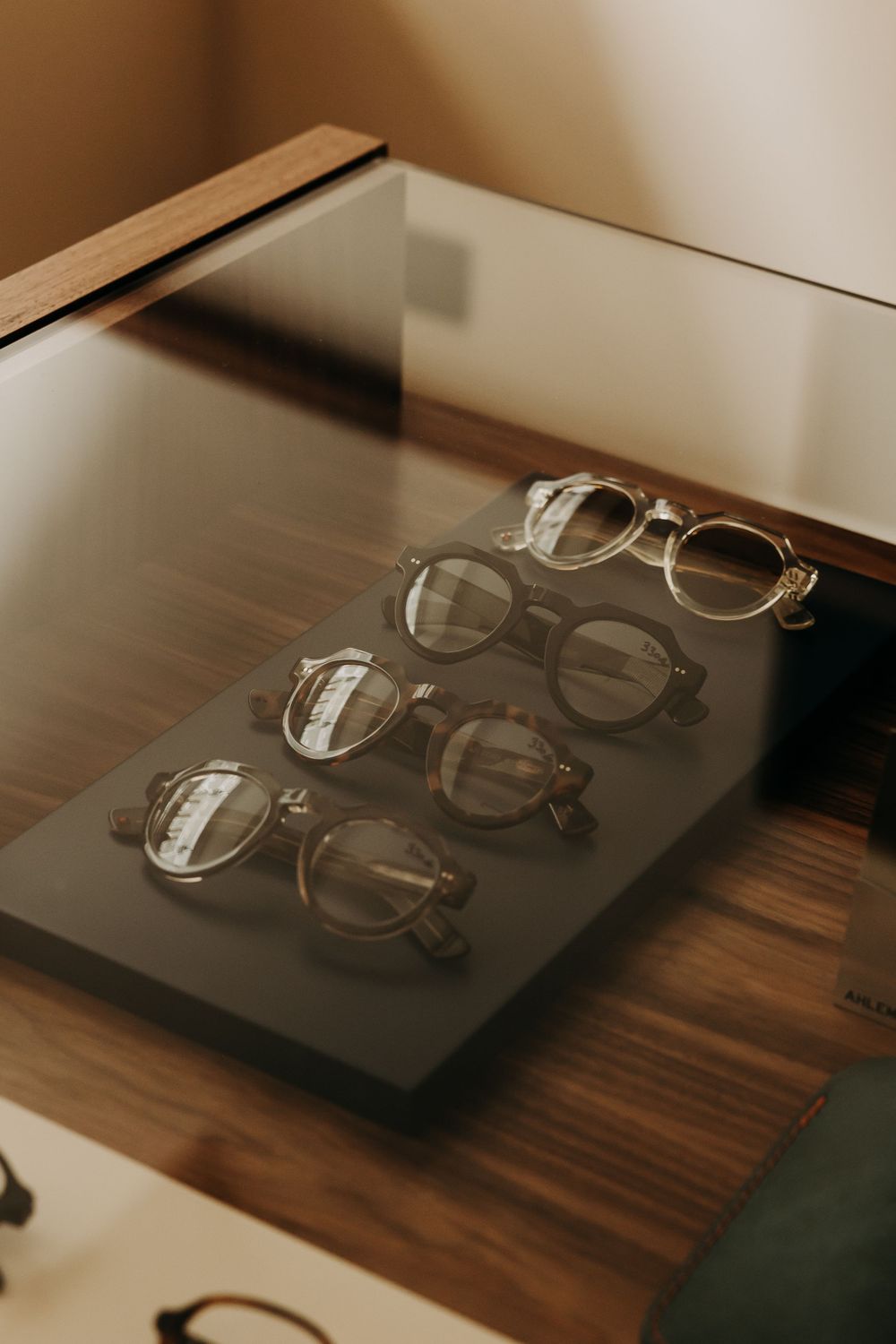 Five stylish eyeglasses frames displayed on a dark tray inside a glass showcase on a wooden table.