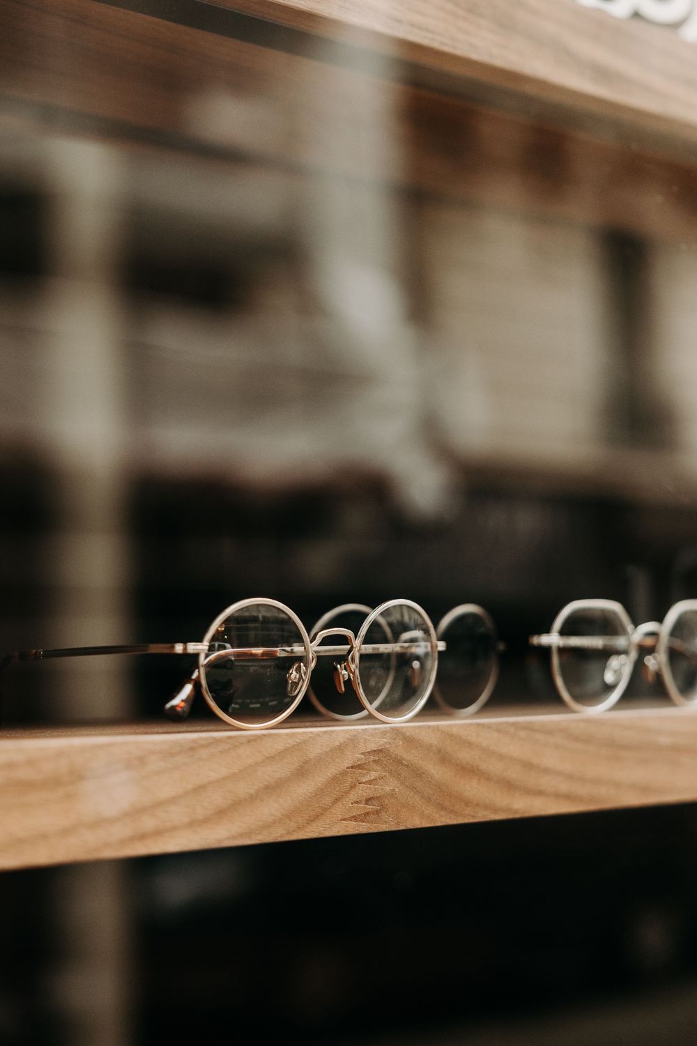 Elegant round eyeglasses displayed on a wooden shelf in an optical store, with a blurred urban reflection in the background.