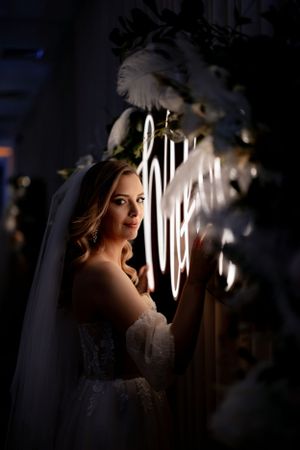 Artistic bridal portrait with dramatic lighting and neon signage behind white floral decorations.