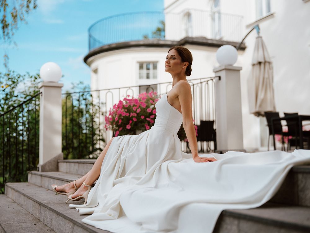 A bride in a flowing white wedding dress sits elegantly on a terrace overlooking a scenic view.