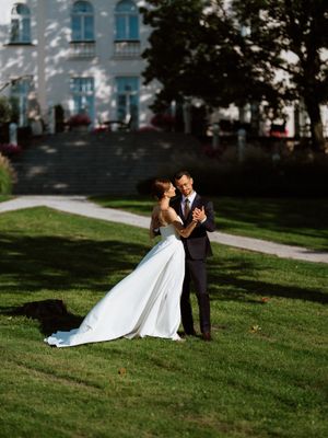 A couple in wedding attire dances together on a sunlit lawn with a white building in the background.