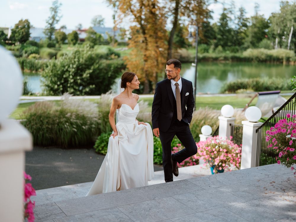 A couple in wedding attire walks up elegant white stairs surrounded by pink flowers and a scenic lake view.