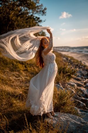 Bride in flowing dress poses dramatically on coastal cliff at sunset.