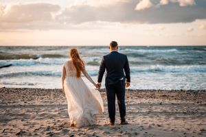Couple holding hands while walking on beach at sunset in wedding attire.