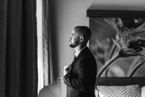 A groom in formal attire adjusts his tie while looking out a window in dramatic lighting.