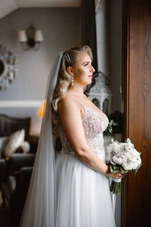 Elegant bride in lace gown holds white bouquet while standing by window in natural light.