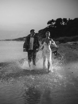 A couple splashes through shallow water on a beach at dusk wearing formal attire in black and white photography.