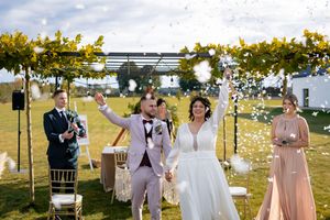 A joyous outdoor wedding celebration under a floral arch with guests standing in a garden setting on a sunny day.