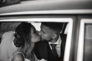 A romantic black and white photo of newlyweds sharing a kiss in a car on their wedding day.