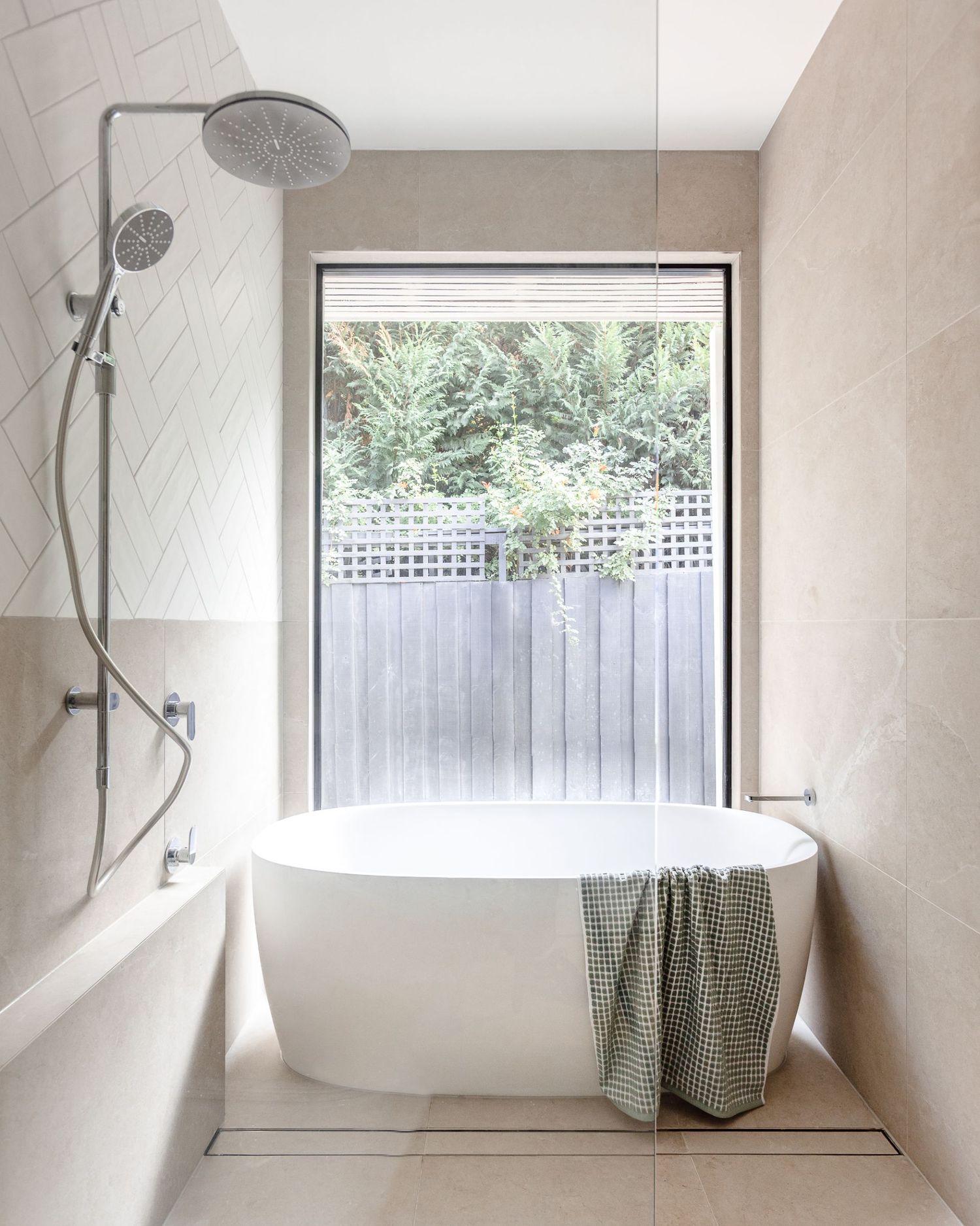 Minimalist bathroom with freestanding white tub and window overlooking garden with privacy screen.