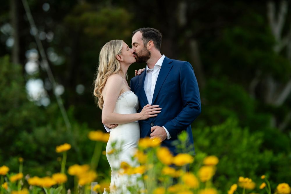 A bride and groom share a romantic kiss surrounded by yellow wildflowers and lush green trees outdoors.