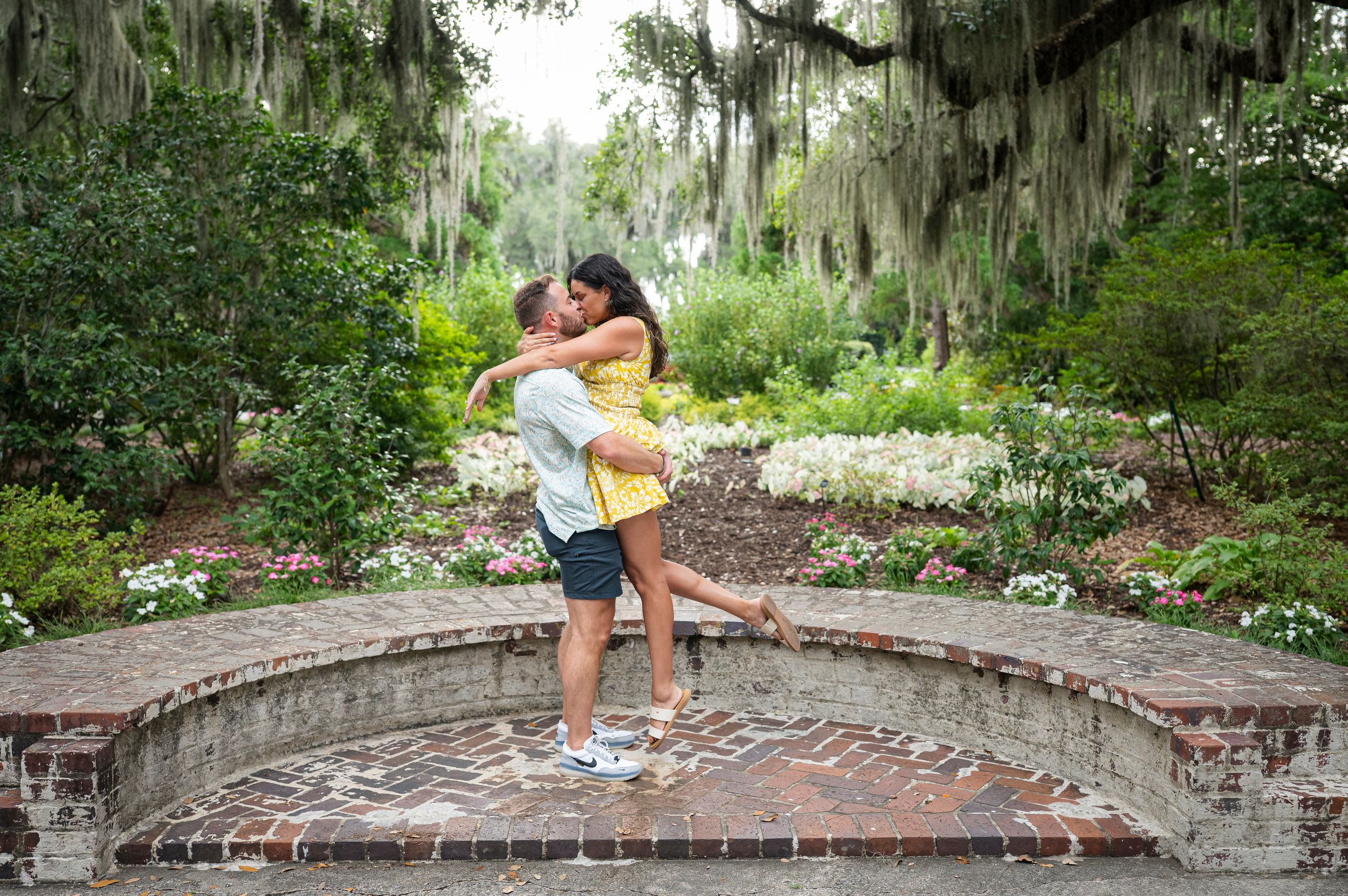 Couple embracing romantically in a lush garden with Spanish moss-draped trees and colorful flowers.