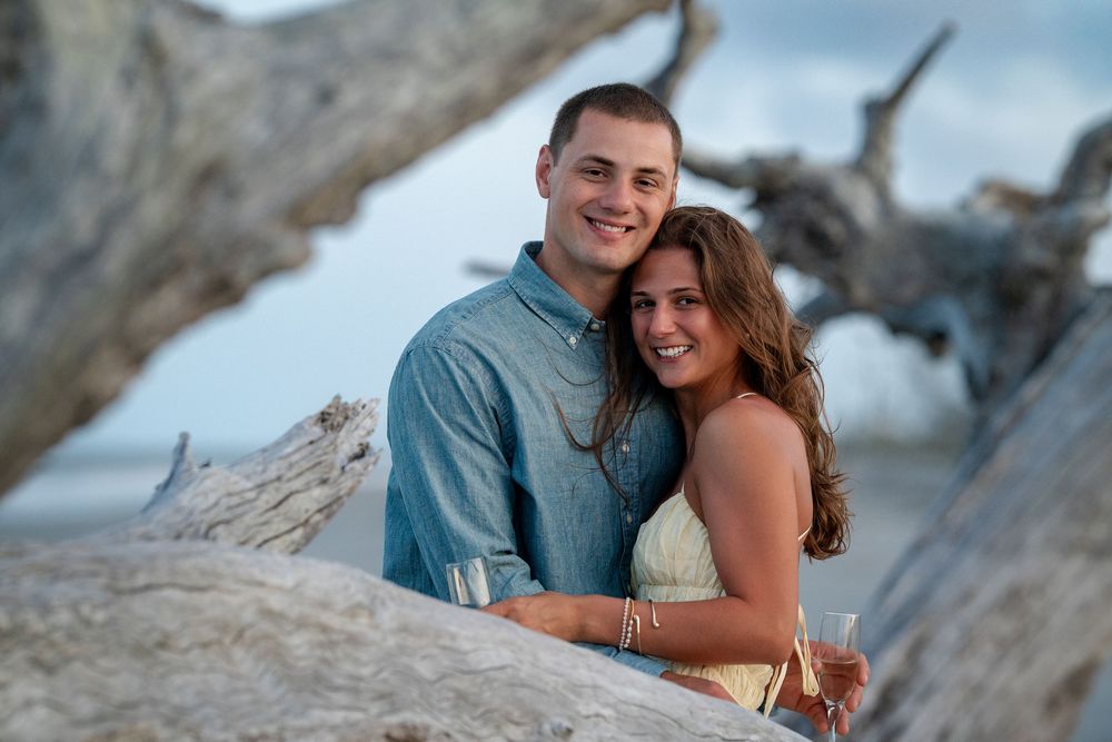 A happy couple embraces while posing between driftwood on the beach at sunset.