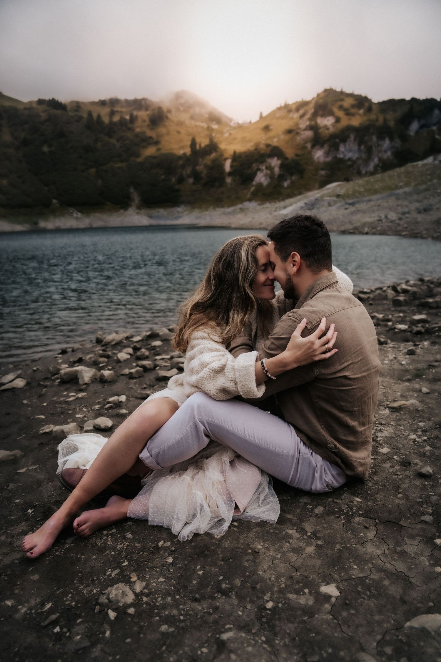A romantic couple in formal attire poses dramatically against a moody forest backdrop with tall evergreen trees.