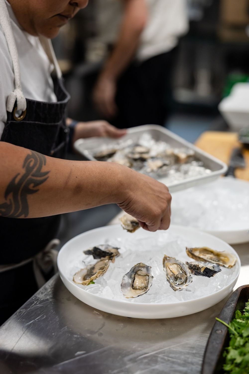 A chef with tattooed arm plates fresh raw oysters on ice at a restaurant prep station.