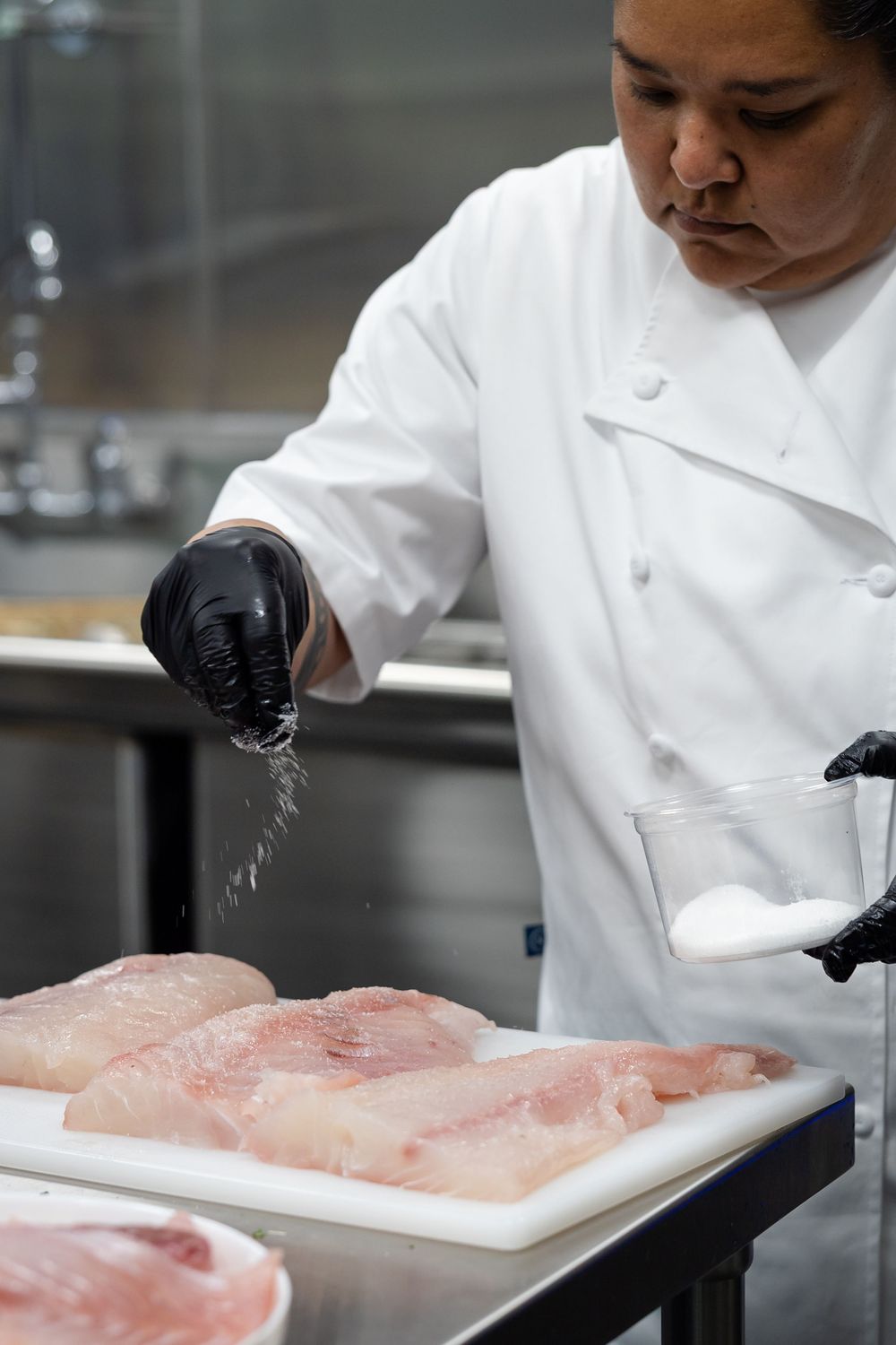Chef in white coat and black gloves prepares raw fish on white cutting board in professional kitchen.