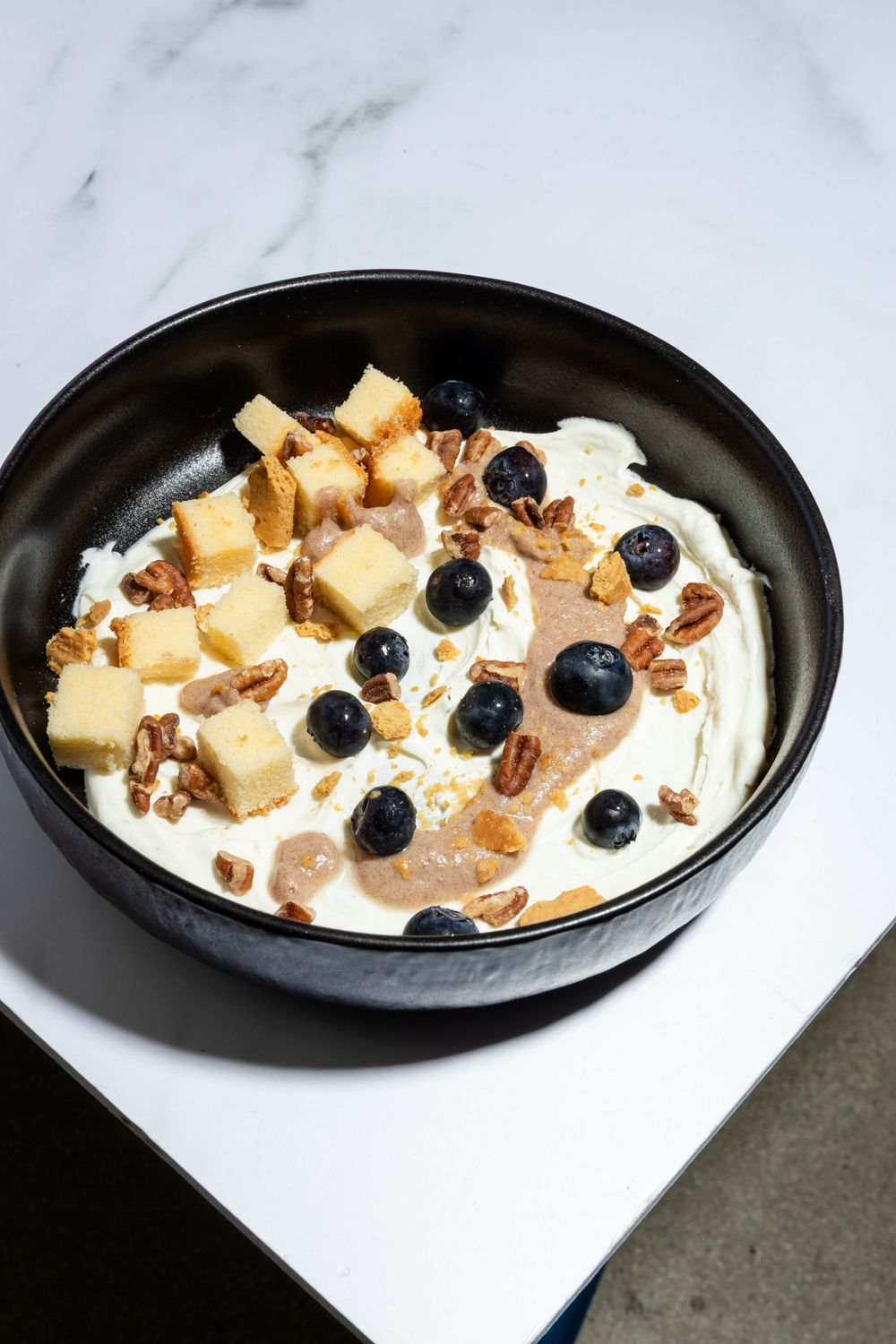 Dessert bowl filled with whipped cream, chopped pecans, fresh blueberries and cubed pound cake on white plate.