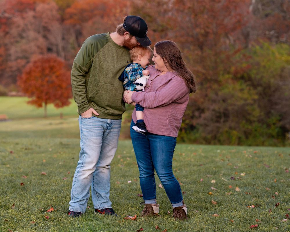 A couple embraces their small child while standing in a grassy field during autumn with orange foliage in background.