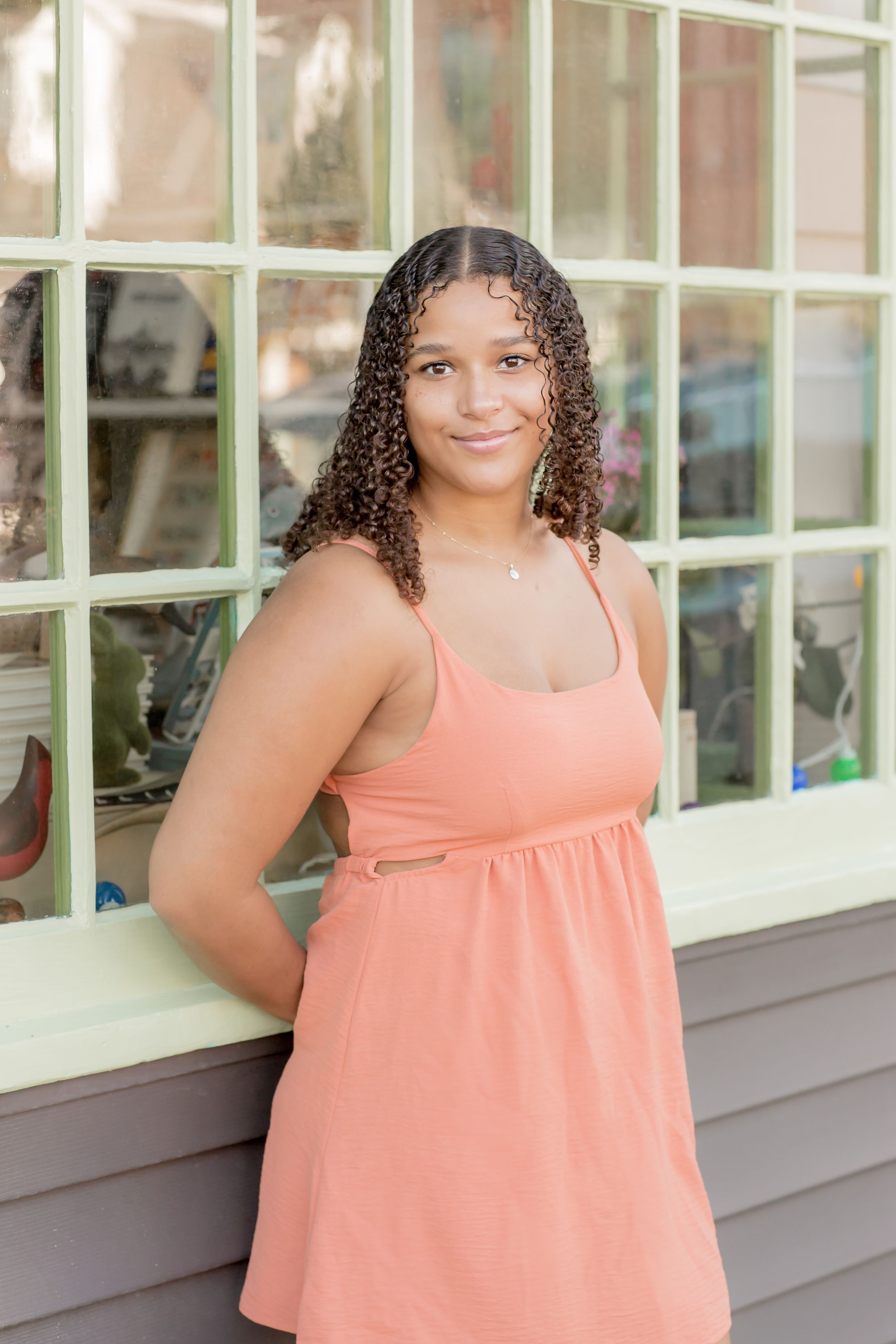 Young woman with curly hair smiling in a peach sundress leaning against a storefront window.