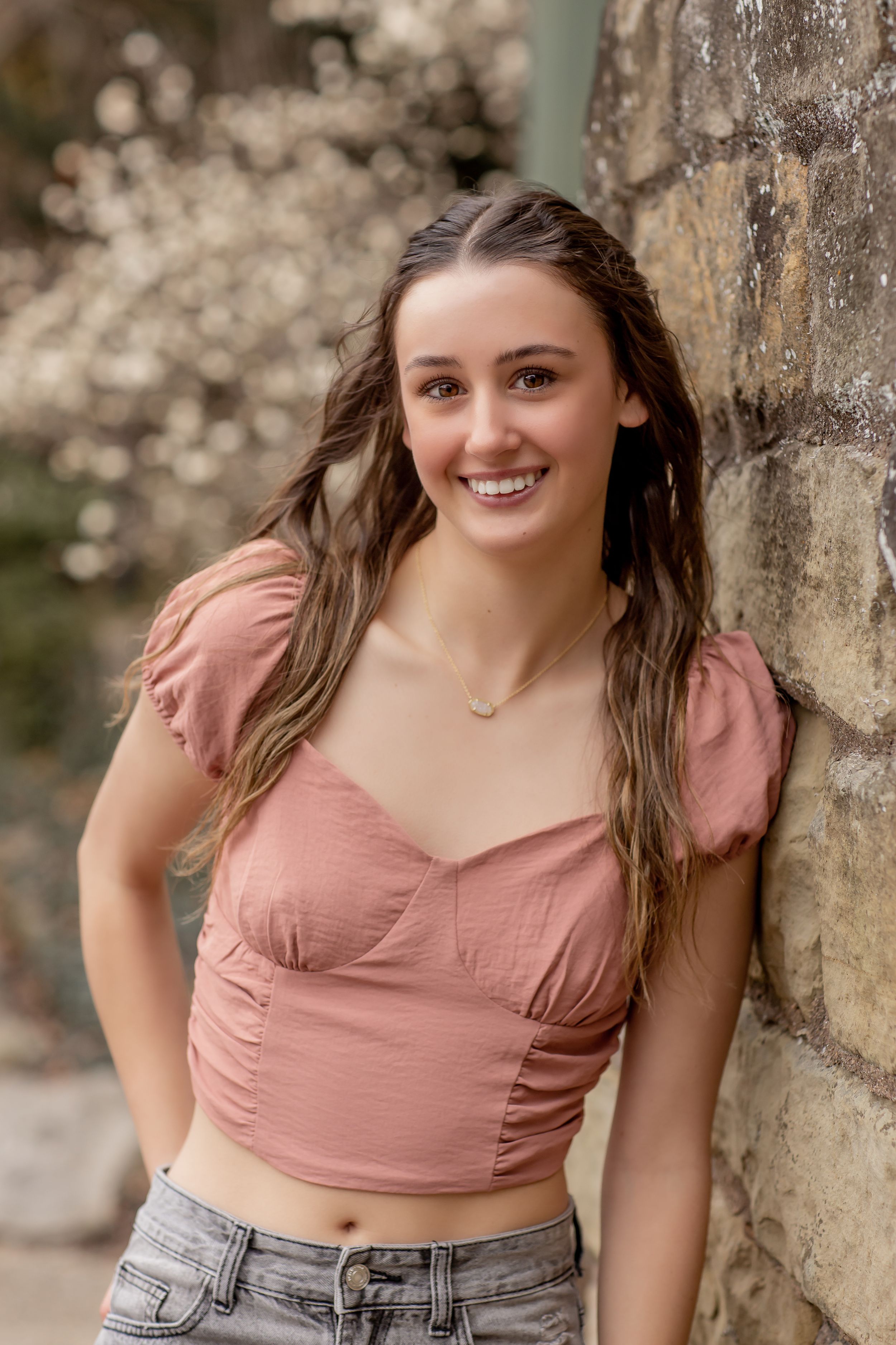 Smiling teen girl with long highlighted hair in a pink crop top leaning against a stone wall outdoors.