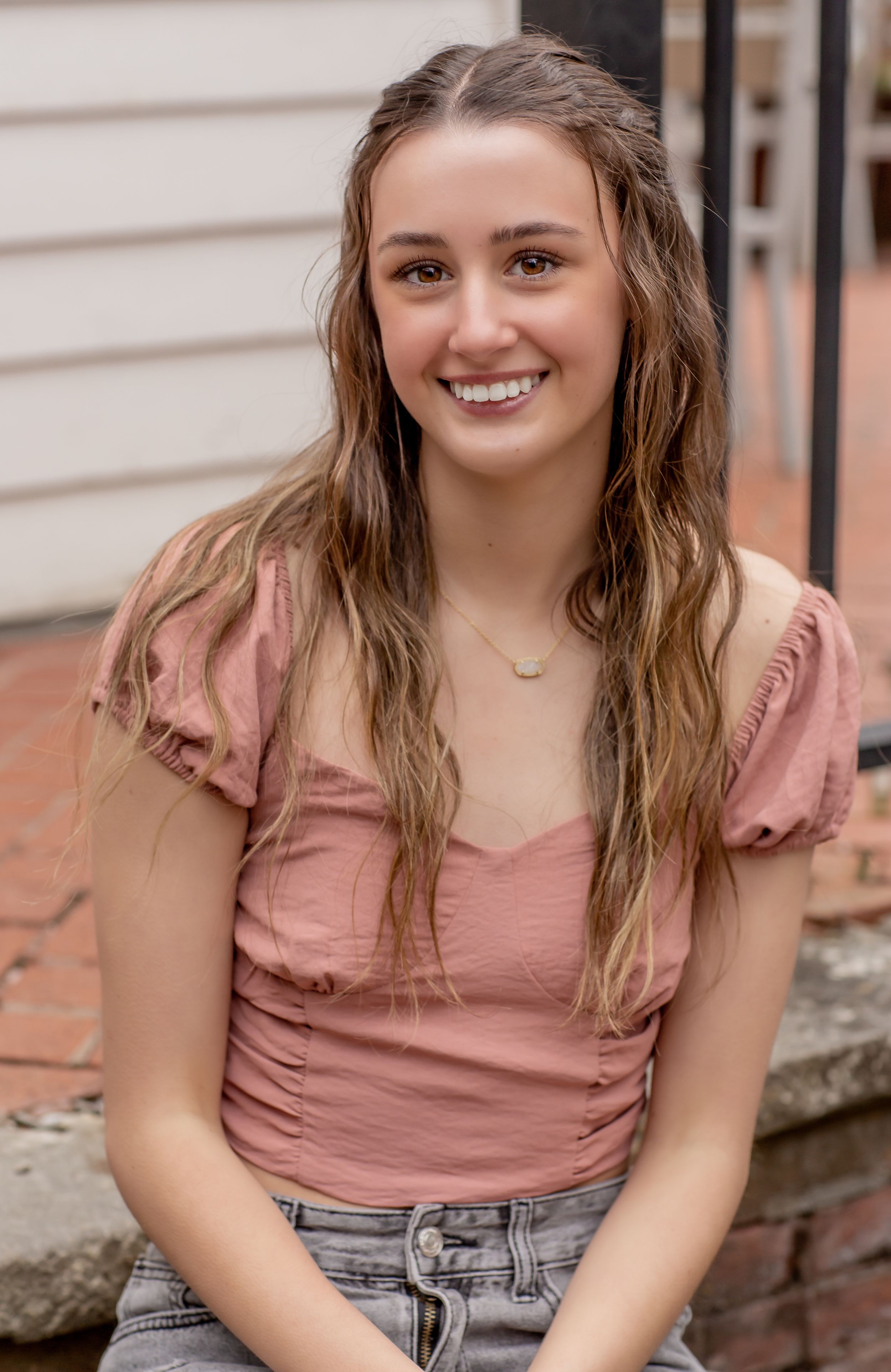 Smiling teenage girl with long wavy highlighted hair wearing a pink top sitting on brick steps outdoors.