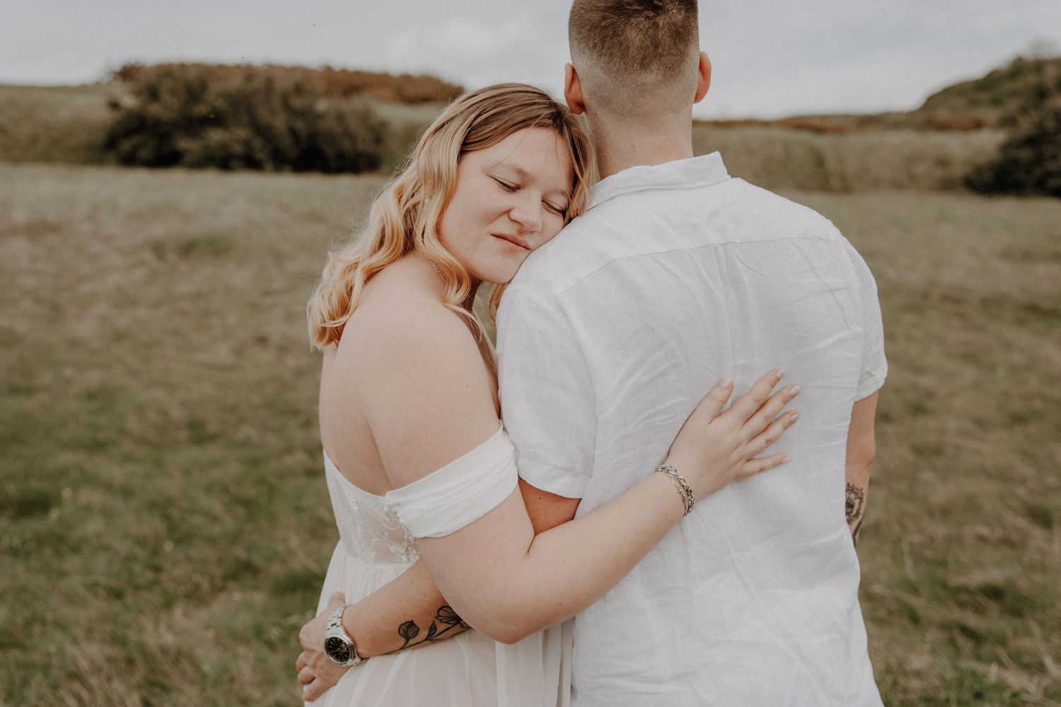 Couple embraces in an intimate moment while standing in a green meadow on an overcast day.