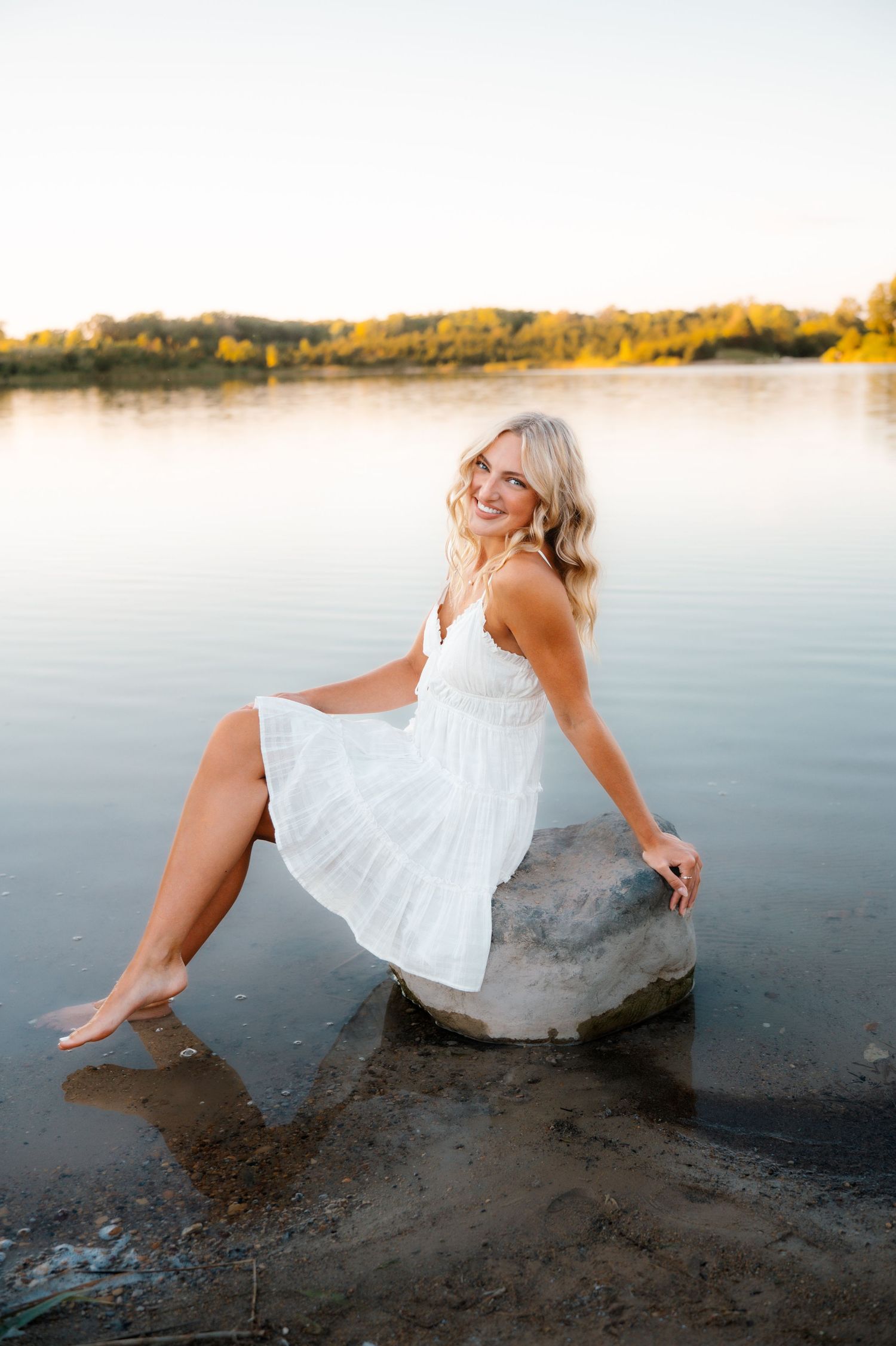Smiling blonde woman in white dress sitting on a rock in a serene lake at sunset.
