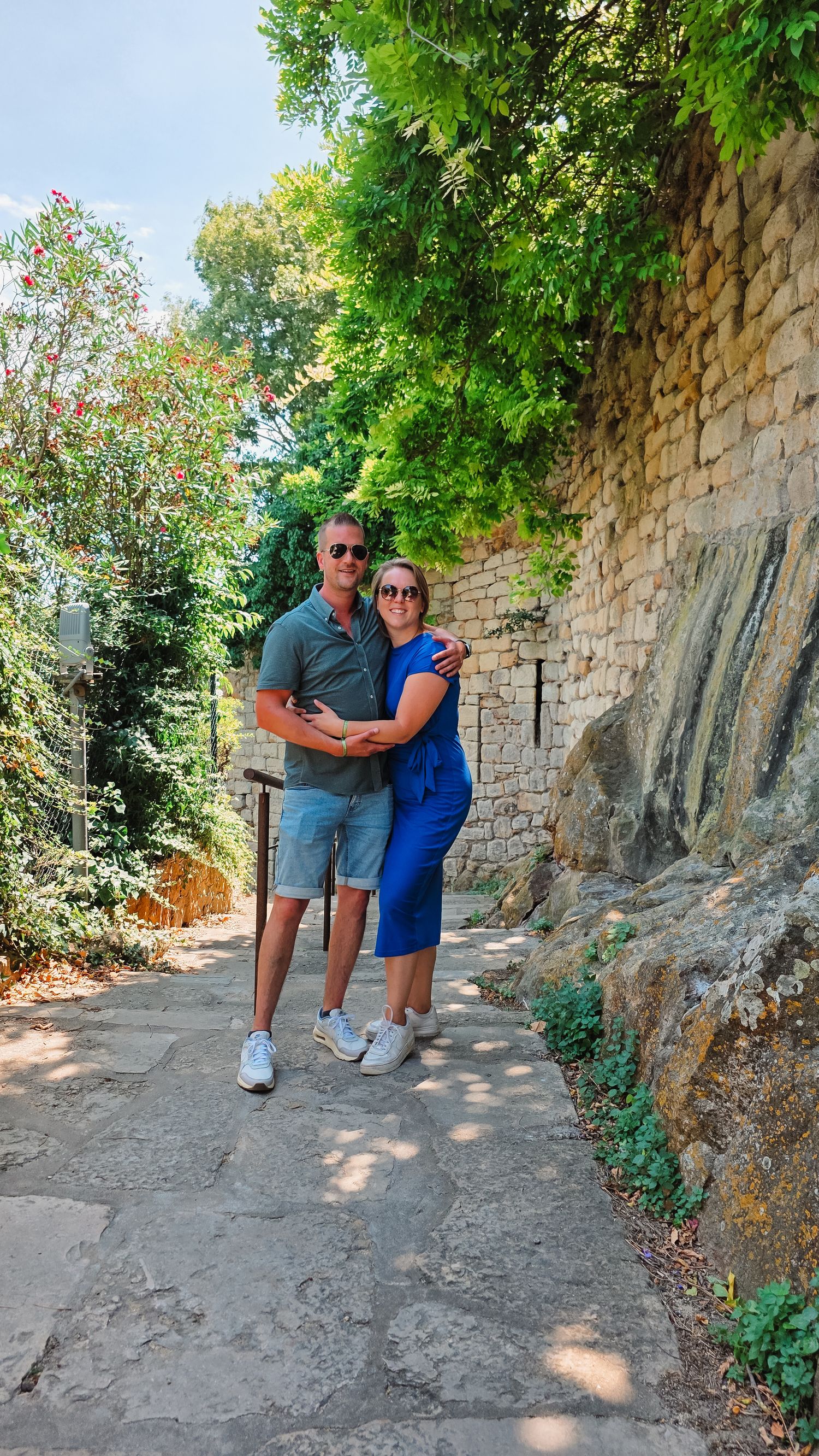A couple walking and posing along a stone wall path surrounded by lush green trees.