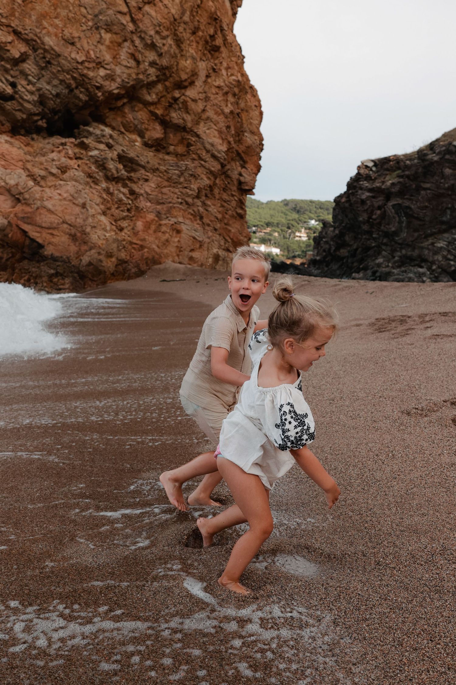 Children playing and splashing in waves near large beach rock formation.