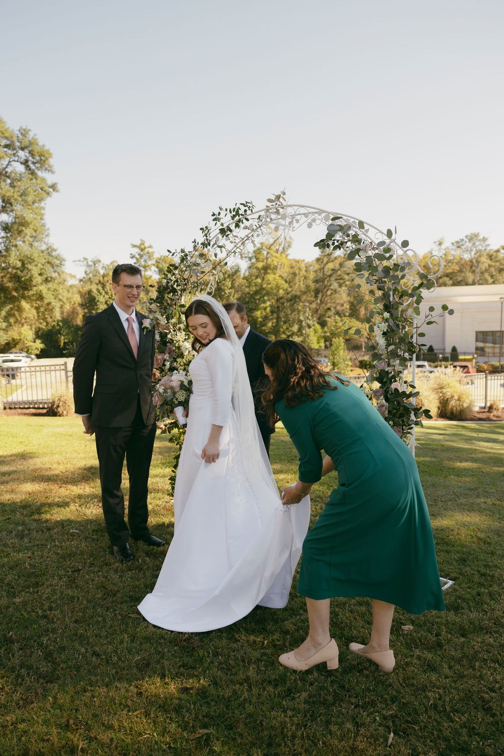 Wedding party members help adjust a bride's dress on a grassy lawn during an outdoor ceremony.