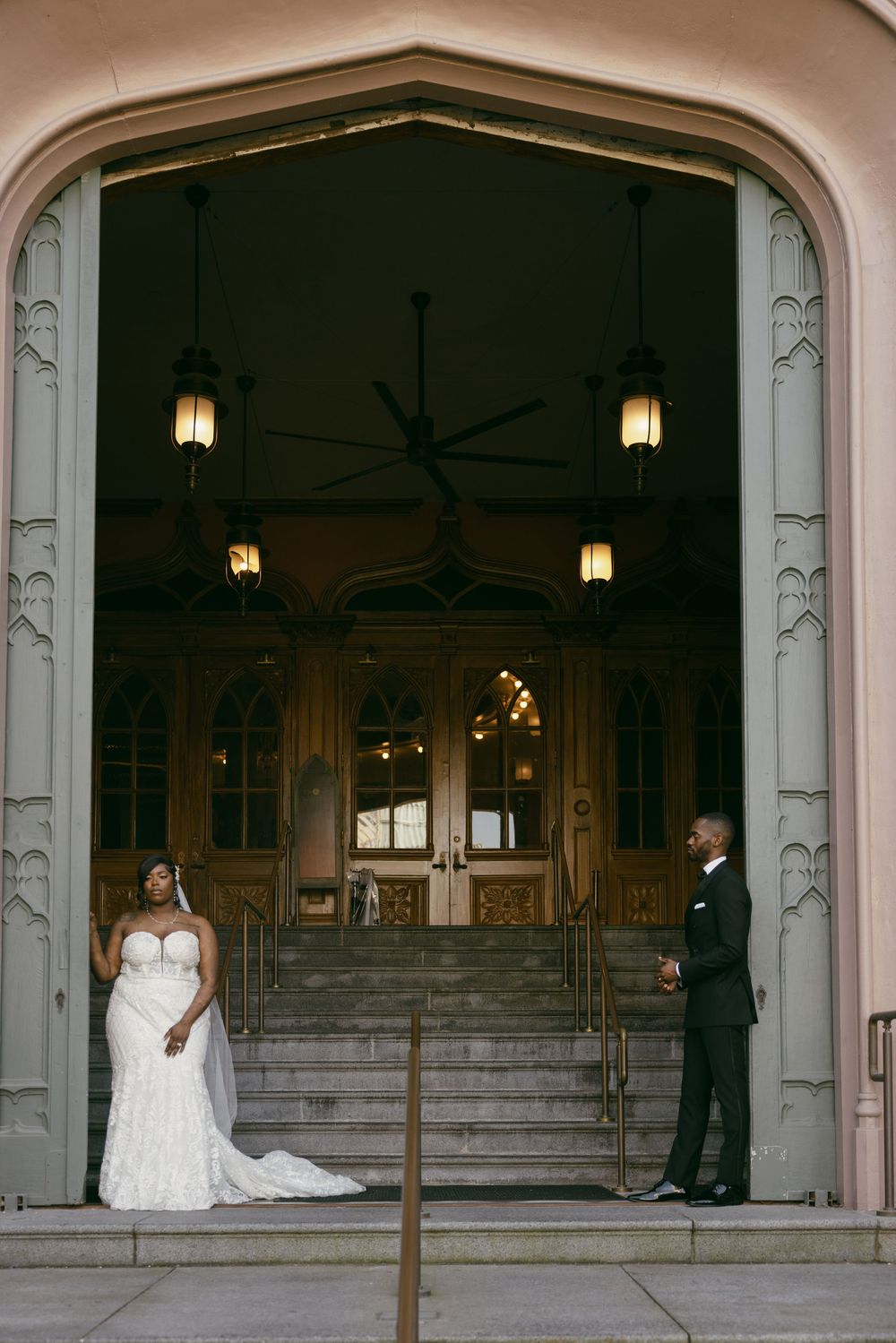 A wedding couple stands apart under elegant stone archways with hanging lanterns in a classic architectural setting.