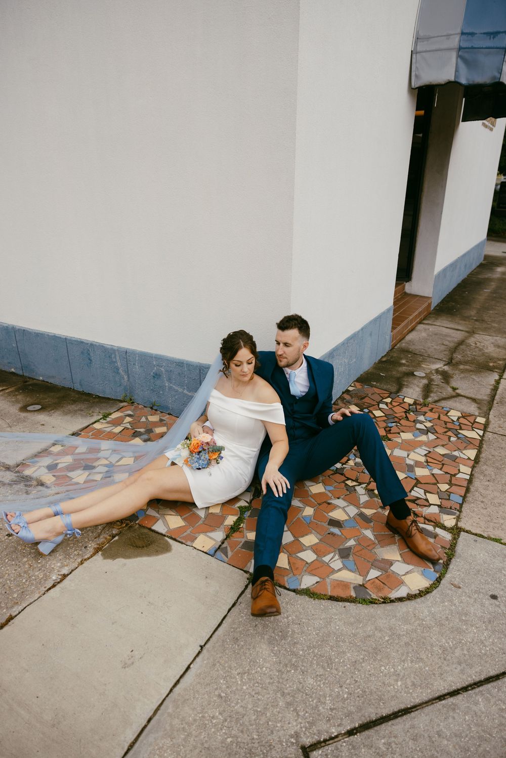 A couple sits on a tiled floor against a white wall in a series of artistic wedding photos.
