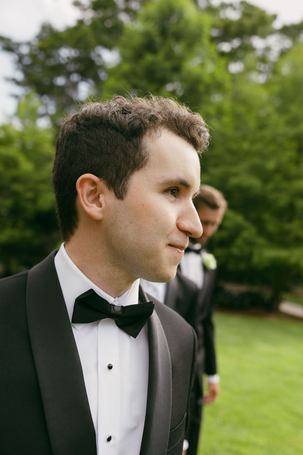 A groom in a black tuxedo and bow tie stands outdoors with a groomsman visible in the background.