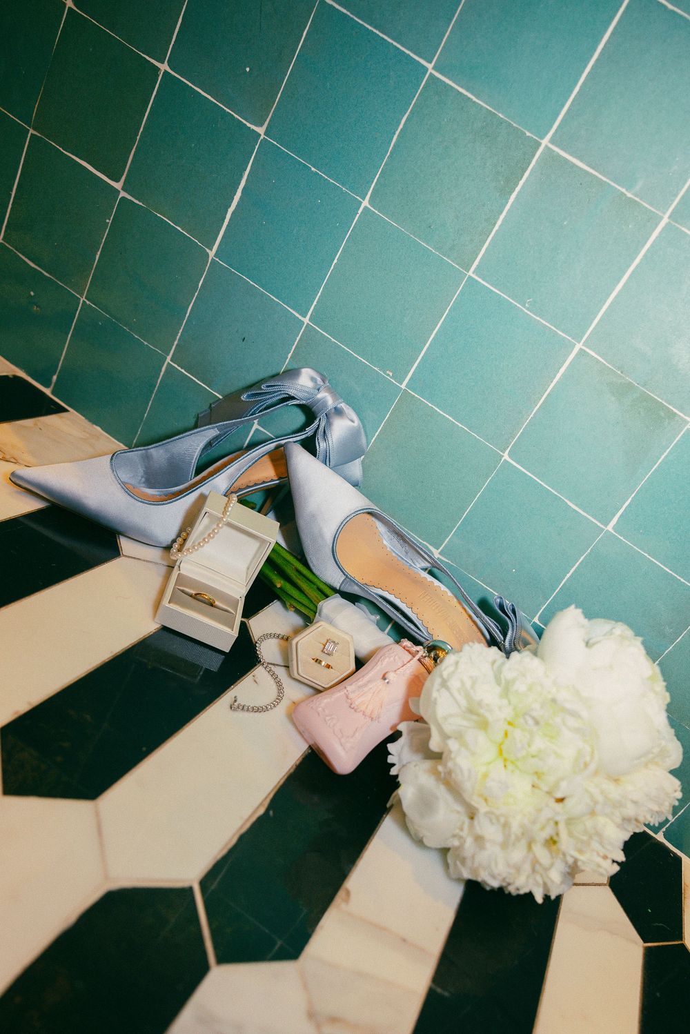 Light blue satin heels, white peony bouquet, and pink accessories arranged on geometric black and white tile floor.