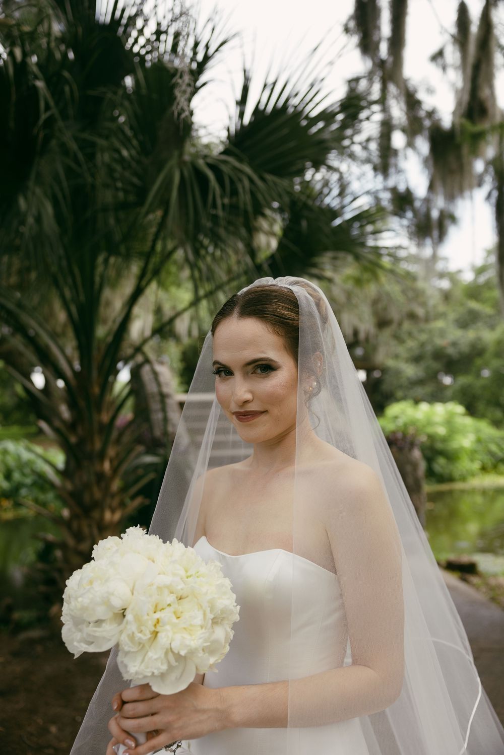 Elegant bride in strapless white gown holding peony bouquet, wearing veil, posing outdoors among lush green tropical foliage.